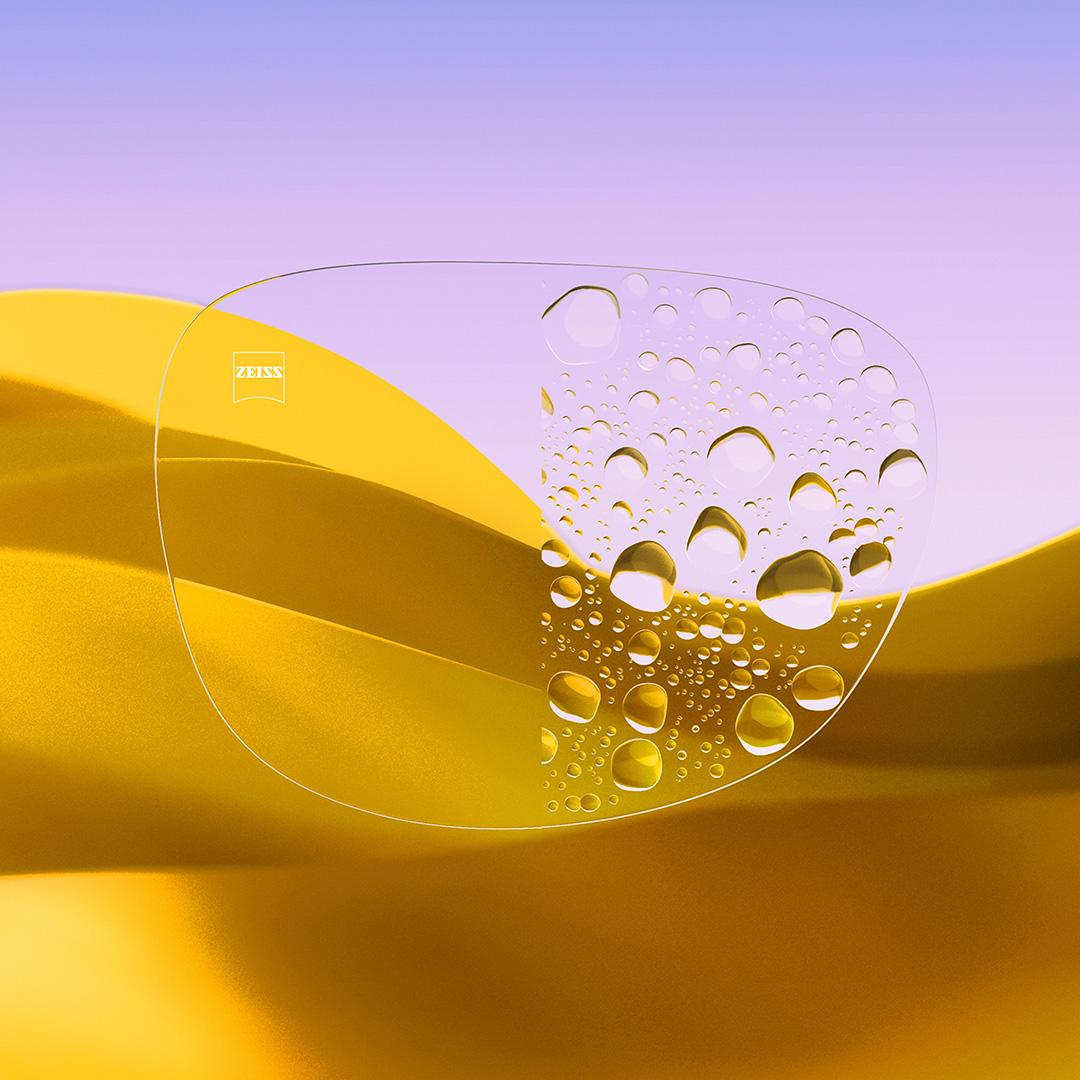 Ett delat glasögonglas där ena sidan, med ZEISS DuraVision Gold UV, är klar och ren, och den andra sidan av glaset är täckt av vattendroppar.