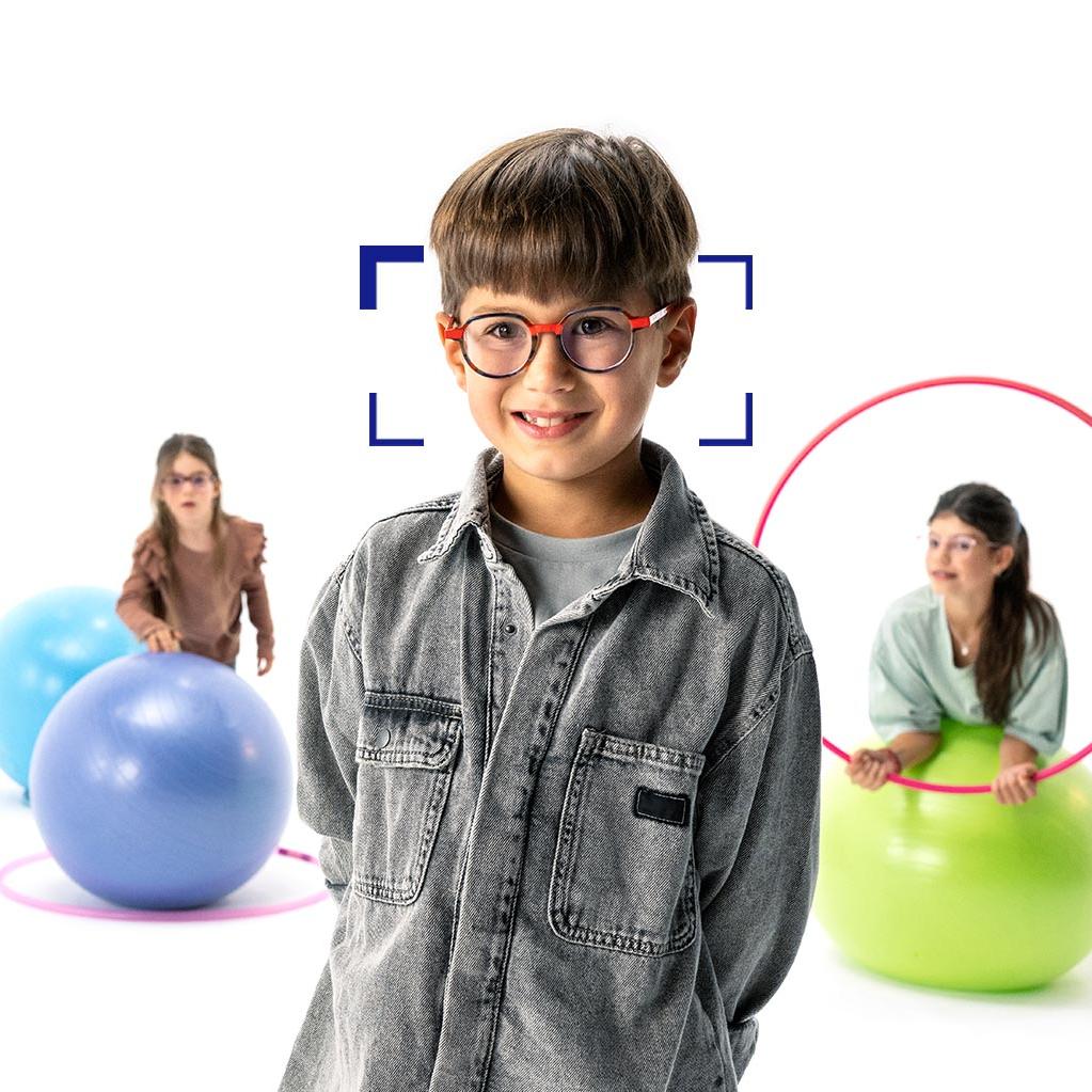 Brunhårig pojke med runda glasögon och ZEISS MyoCare-glas står i förgrunden och ler mot kameran. I bakgrunden leker två flickor, som bär ZEISS MyoCare-glas, med rockringar och pilatesbollar.
