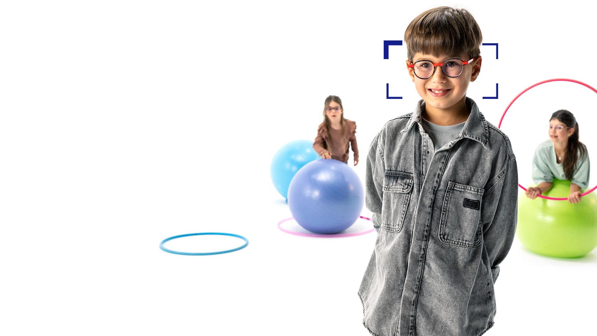 Brunhårig pojke med runda glasögon och ZEISS MyoCare-glas står i förgrunden och ler mot kameran. I bakgrunden leker två flickor, som bär ZEISS MyoCare-glas, med rockringar och pilatesbollar.