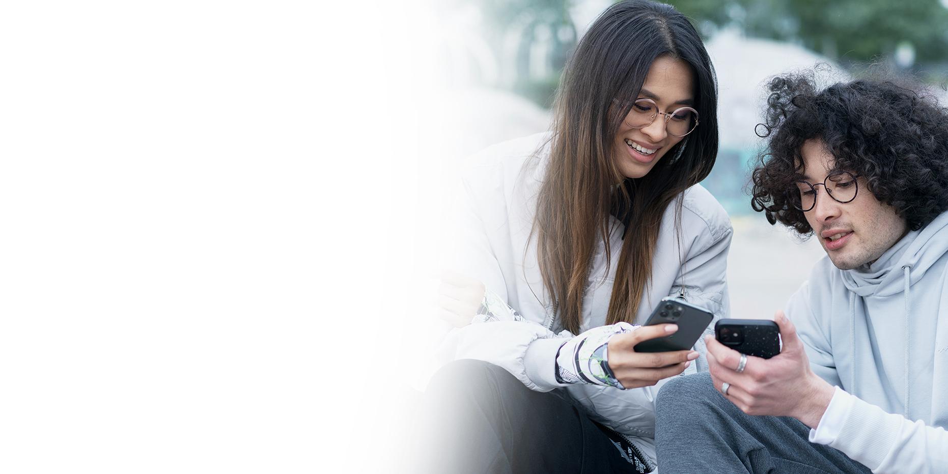 Två unga människor, en kvinna med långt, brunt hår som ler och en pojke med lockigt, svart hår som har på sig ZEISS SmartLife-glasögon tittar på sina telefoner. 