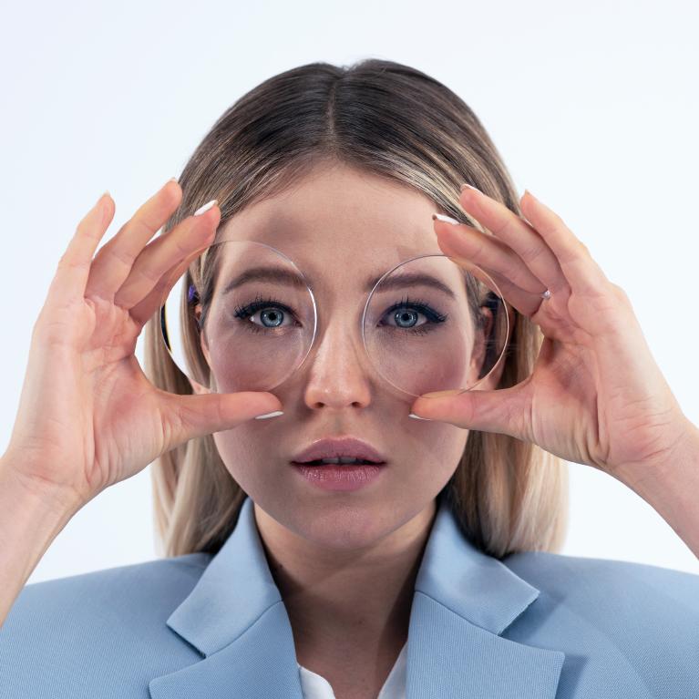En ung blond kvinna håller upp två glas framför ögonen, för att visa hur ögonen tycks förminskas genom tjocka minus-glasögon.