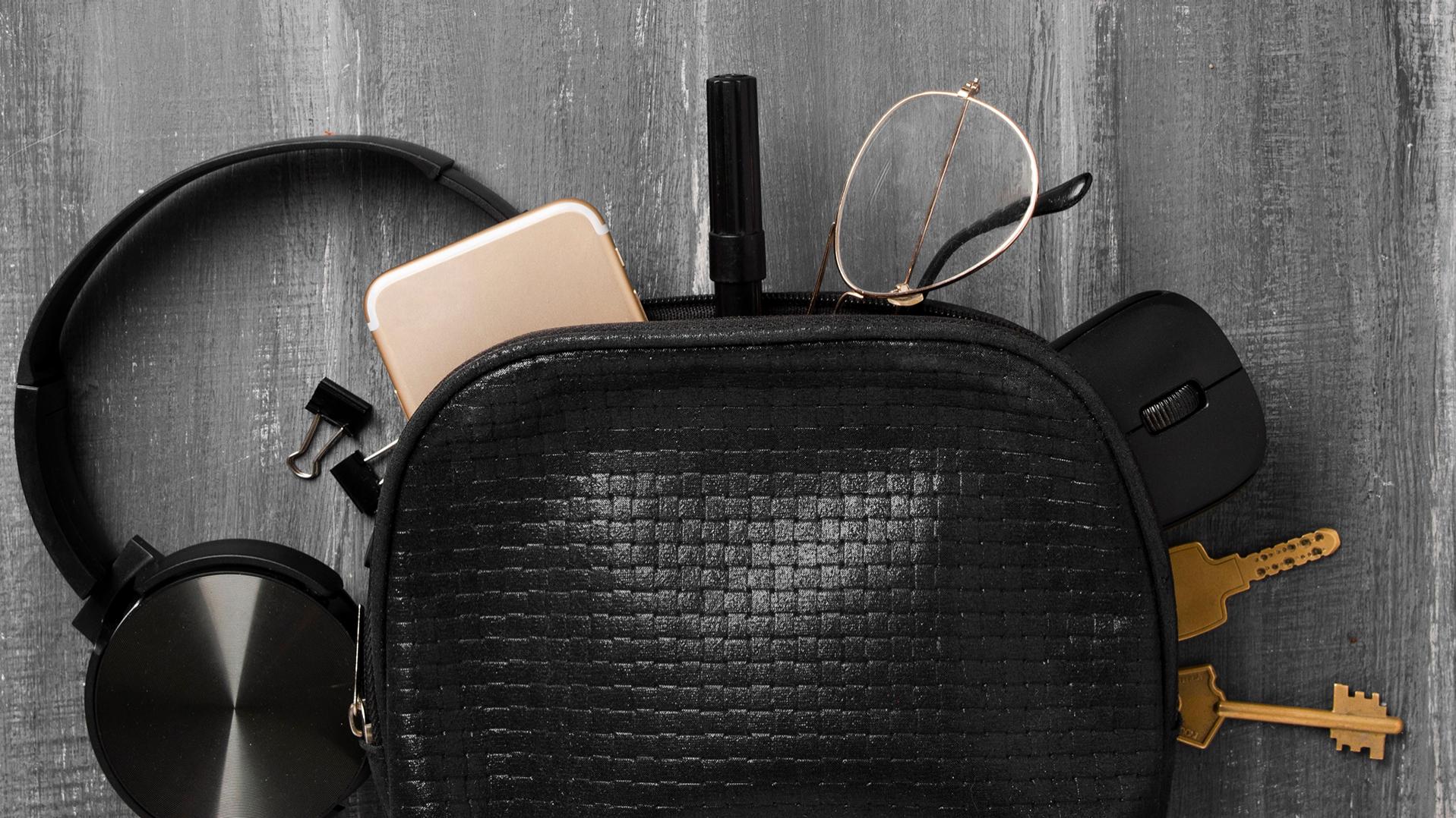En liten svart väska där hörlurar, mobiltelefon, nycklar, pappersklämmor, en penna, en datormus och glasögon med ZEISS-glas försedda med DuraVision-ytbehandling kikar fram och ligger till hälften på det grå golvet.