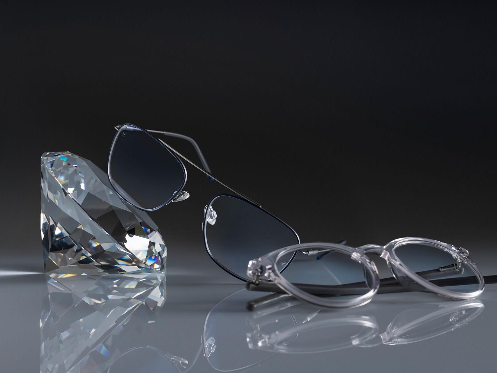 Två par glasögon med ZEISS-glas försedda med ZEISS hårdaste ytbehandling någonsin – DuraVision® Platinum. Ett par glasögon ligger mot en kristall, det andra plant på underlaget. Båda har klara glas utan blåaktiga reflexer.