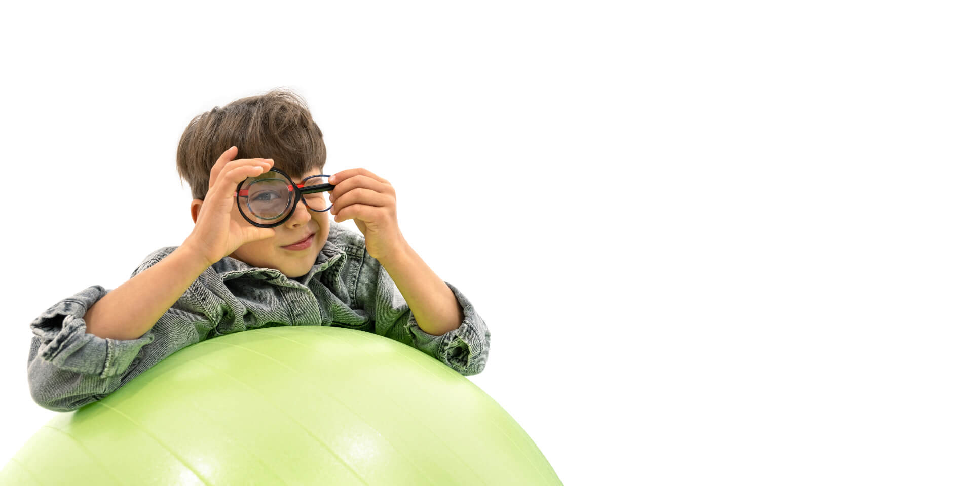 En pojke med ZEISS glas för myopihantering lutar sig mot en gymnastikboll och håller ett förstoringsglas framför ena ögat.