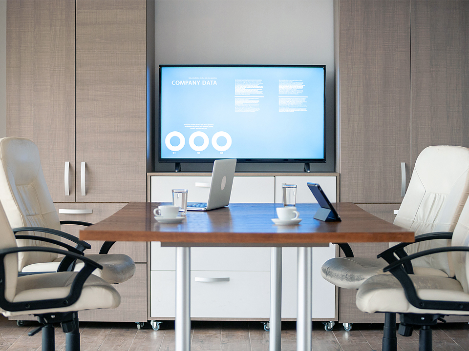 Vy från skrivbordet på chefens kontor. En monitor är uppsatt på väggen några meter bort. Det finns ett litet mötesbord med kontorsstolar och bärbara datorer på bordet.