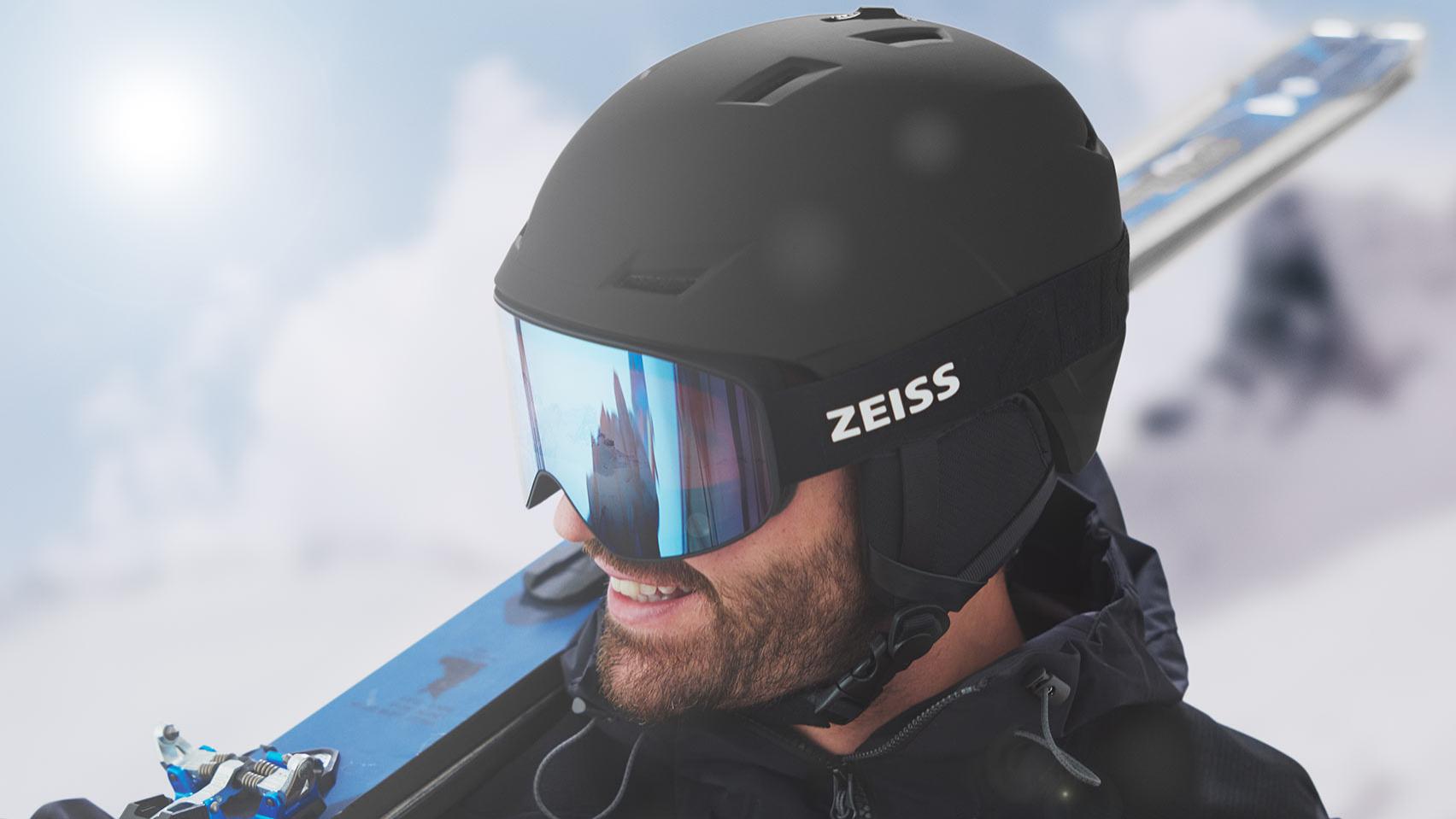 En man bär en svart hjälm med ZEISS skidglasögon med blå spegellinser, och har sina skidor över höger axel.