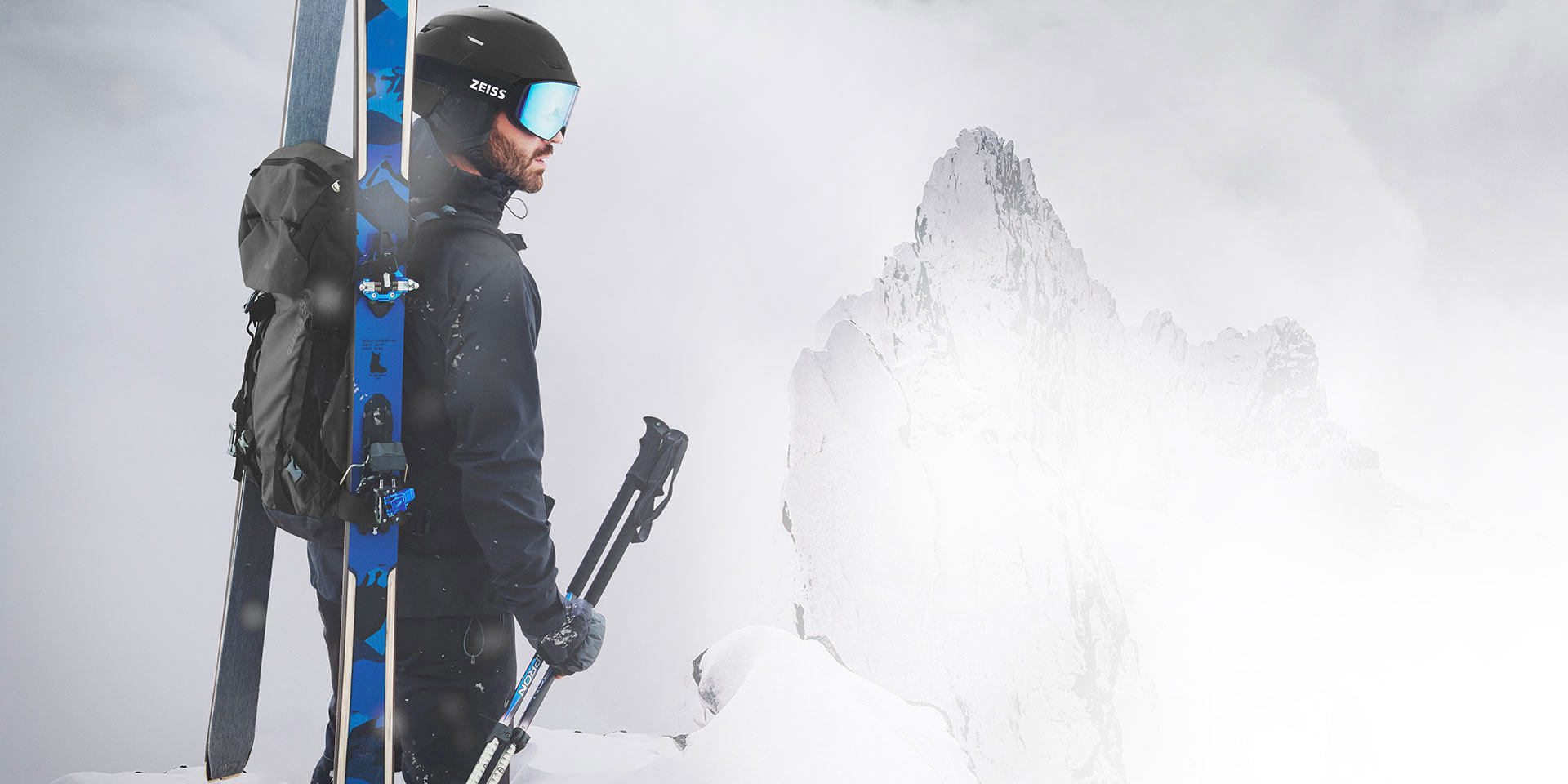 En man på skidor bär en svart hjälm med ZEISS skidglasögon med blå spegellinser, har en ryggsäck på ryggen och håller stavarna i höger hand. Han står på en bergstopp.