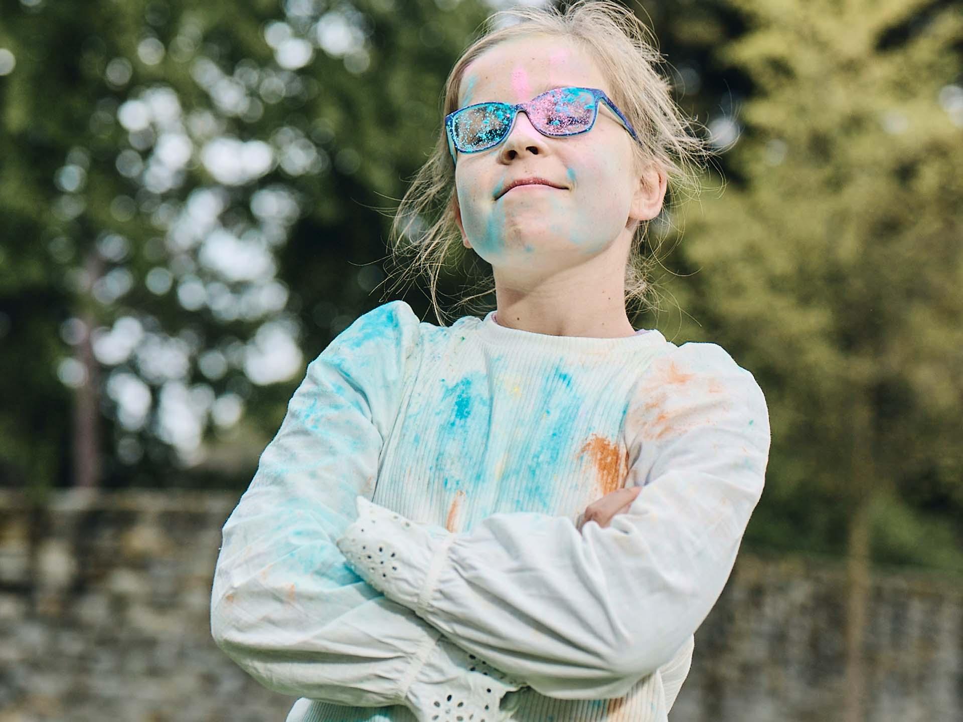 En flicka med armarna i kors och smutsiga glasögon efter att ha lekt med färgpulver tittar finurligt och ler.