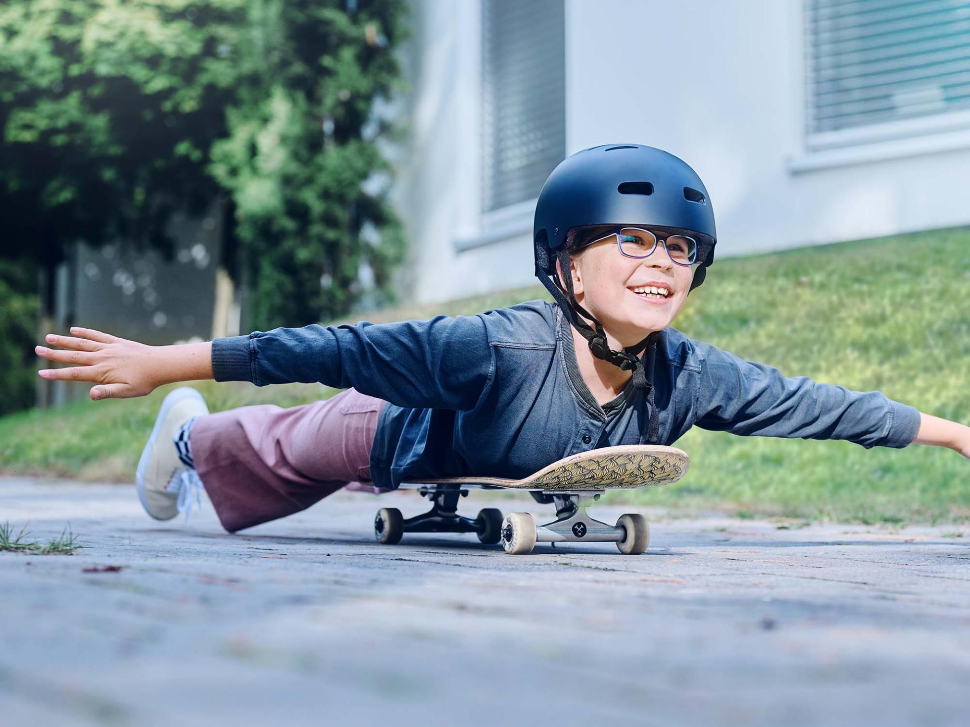 En flicka med hjälm och glasögon rullar ner för gatan liggandes på en skateboard och sträcker ut armarna.