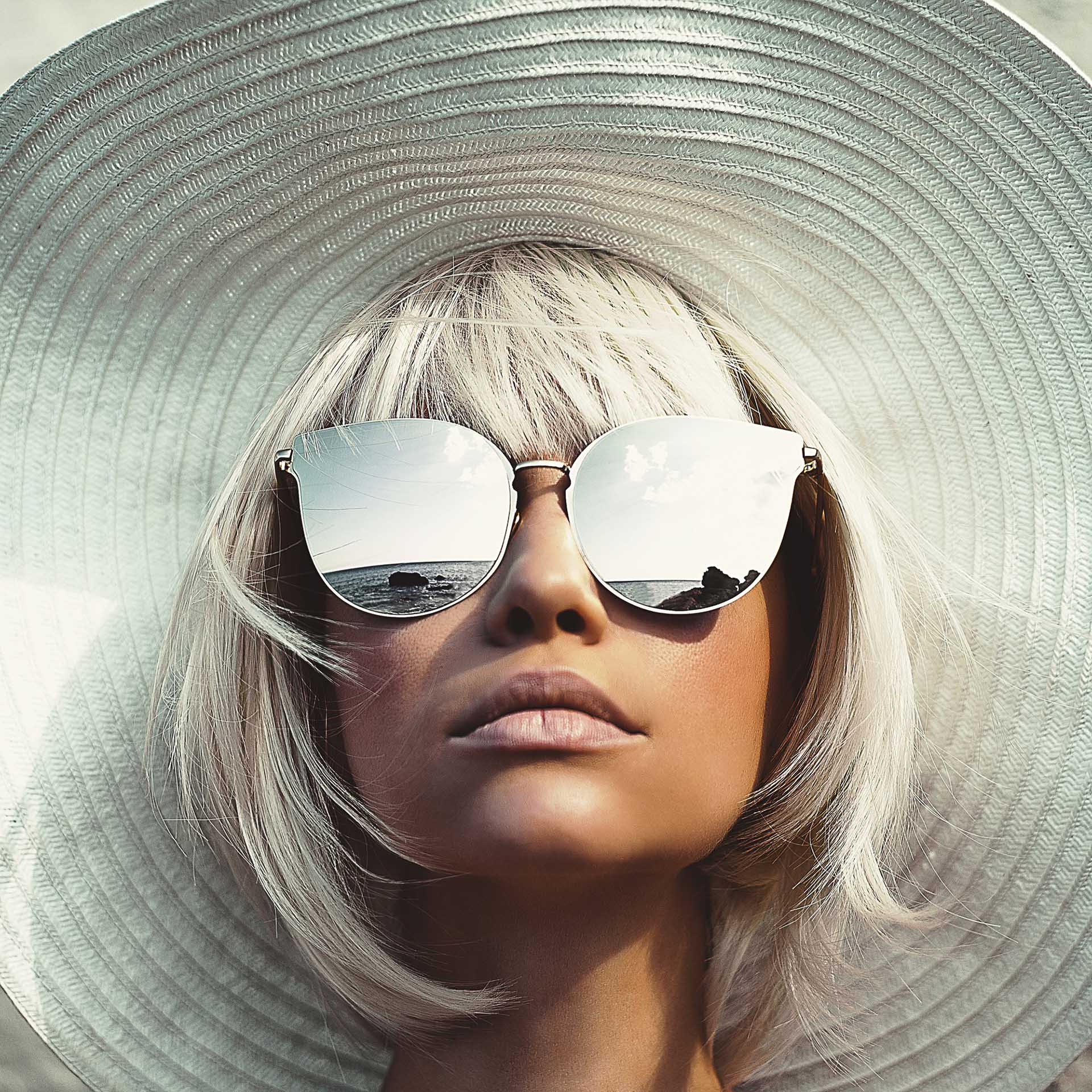 Ung kvinna med en stor solhatt tittar upp, och bär stora solglasögon med spegeleffekt.
